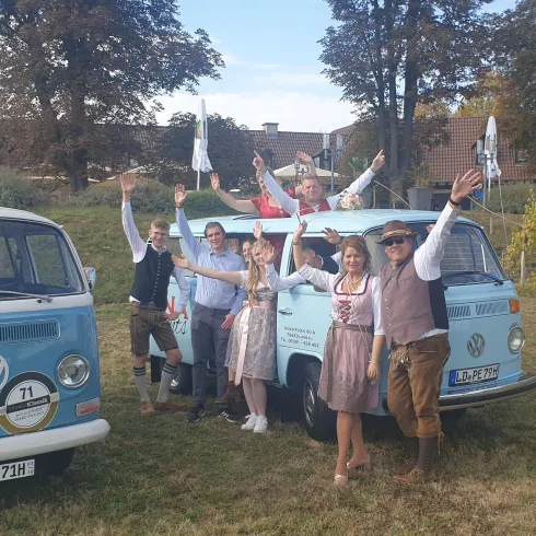 Eine Gruppe von Menschen steht fröhlich neben zwei blauen Volkswagen Bullis am Deutschen Weintor. Im Hintergrund sind Bäume und Weinreben zu sehen.