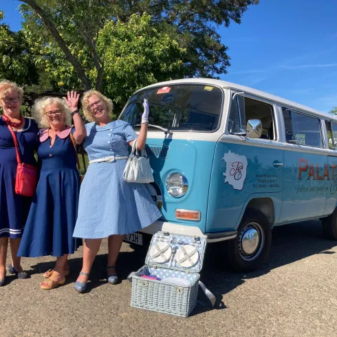 Drei Frauen in bunten Kleidern stehen neben einem blauen VW-Bus und winken freundlich. Im Hintergrund sind Bäume und eine sonnige Landschaft zu sehen.