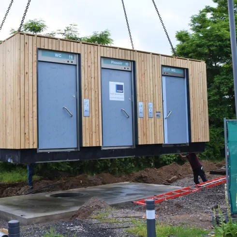 Ein moderner Baucontainer wird mit einem Kran angehoben. Im Hintergrund sind Bäume und eine Baustelle zu sehen.