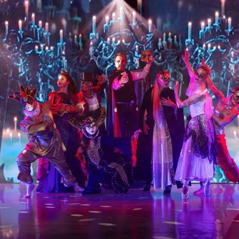 Eine dramatische Aufführung mit Tänzern in bunten Kostümen und Masken. Im Hintergrund leuchtet eine geheimnisvolle Kulisse mit Kerzen und Schatten.