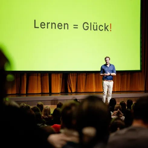 Ein Redner steht auf einer Bühne vor einem großen, grünen Bildschirm mit der Aufschrift „Lernen = Glück!“. Im Publikum sitzen viele Menschen und hören aufmerksam zu.