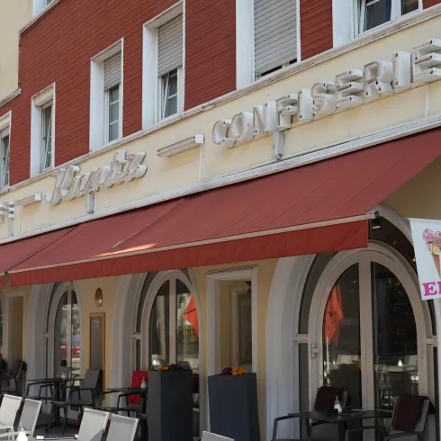 Ein gemütliches Café mit einem roten Markisen und einem Schild für Eis. Die Fassade ist hell und einladend, mit mehreren Stühlen im Freien.