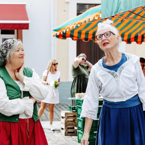 Zwei Frauen in traditionellen Kostümen stehen auf einem Markt und lachen. Im Hintergrund sind Stände mit frischem Obst und Gemüse zu sehen.