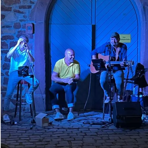 Drei Musiker stehen vor einer blauen Tür und spielen live Musik. Es ist eine fröhliche Atmosphäre mit buntem Licht.
