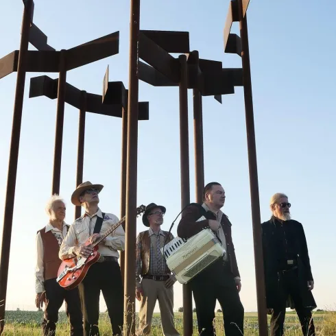 Eine Band steht unter einem Kunstwerk aus rostetem Metall. Sie spielen Instrumente und posieren in einer grünen Wiese.