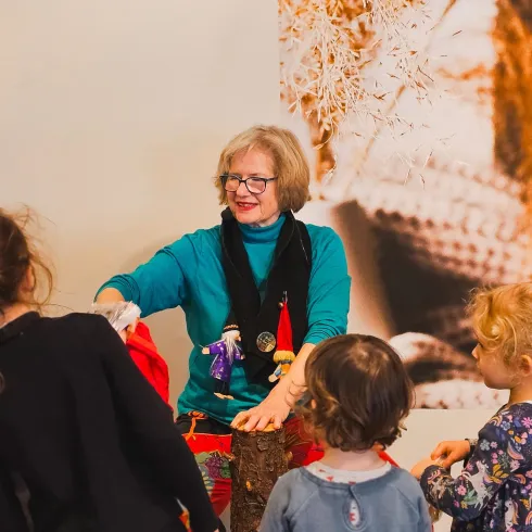 Eine Frau zeigt Kindern, wie man mit Puppen spielt. Sie stehen um sie herum und hören aufmerksam zu.