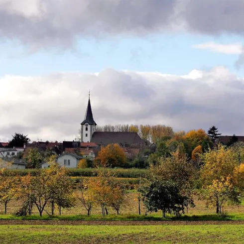 Eine malerische Landschaft mit einem kleinen Dorf im Hintergrund. Die Kirche mit ihrem hohen Turm und herbstlichen Bäumen prägen das Bild.