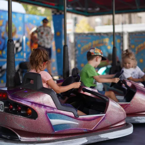 Kinder fahren mit Autoscootern in einem Vergnügungspark. Im Hintergrund sind bunte Dekorationen zu sehen.