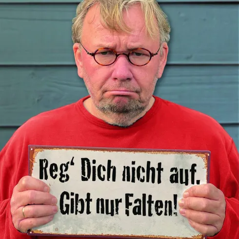 Ein trauriger Mann hält ein Schild in den Händen. Darauf steht: "Reg' Dich nicht auf. Gibt nur Falten!"