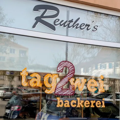 Ein freundliches Bäckerei-Schild mit dem Namen "Reuther's tag2wei backerei". Die Fenster reflektieren die Umgebung und laden Passanten ein.