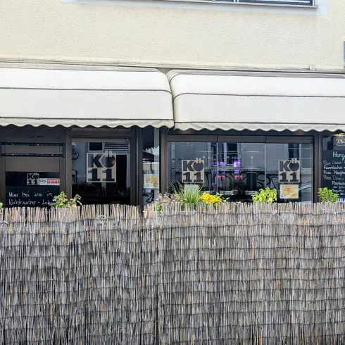 Ein gemütliches Restaurant mit einer Markise und einem Sichtschutz aus Holz. Vor dem Gebäude sind Pflanzen und eine einladende Atmosphäre zu sehen.