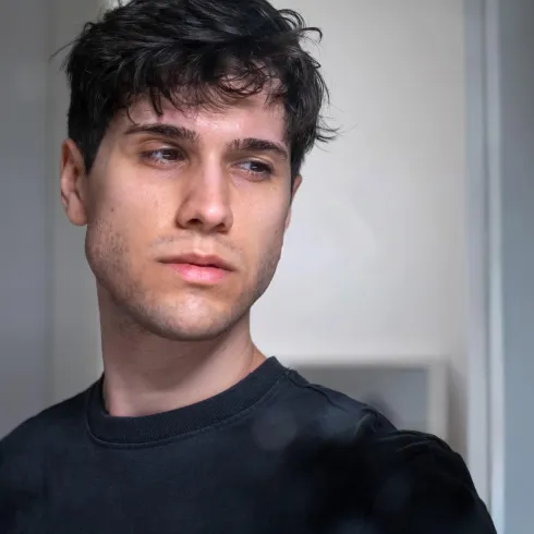 Ein junger Mann mit dunklen, lockigen Haaren blickt nachdenklich zur Seite. Er trägt ein einfaches schwarzes Shirt und der Hintergrund ist unscharf.