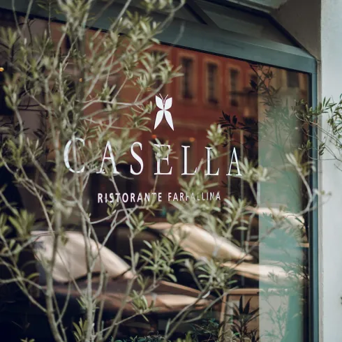 Ein Fenster mit dem Logo des Restaurants Casella. Im Vordergrund sind grüne Pflanzen zu sehen, die eine angenehme Atmosphäre schaffen.