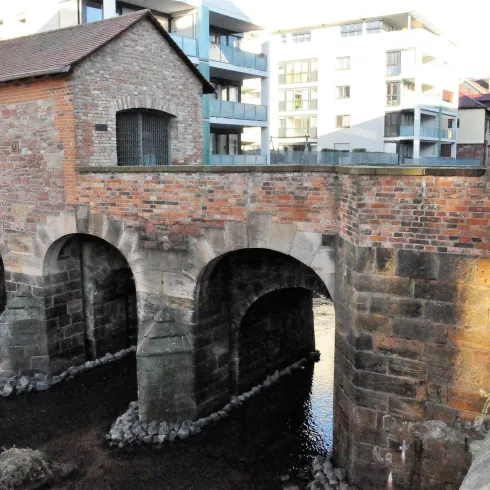 Eine alte Steinbrücke mit einem kleinen Gebäude auf der linken Seite. Im Hintergrund sind moderne Wohngebäude zu sehen.