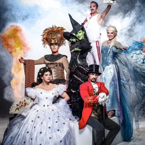 Eine Gruppe von Menschen in festlichen Kostümen steht vor einem dramatischen Hintergrund. Die Charaktere scheinen aus verschiedenen berühmten Geschichten oder Musicals zu stammen.