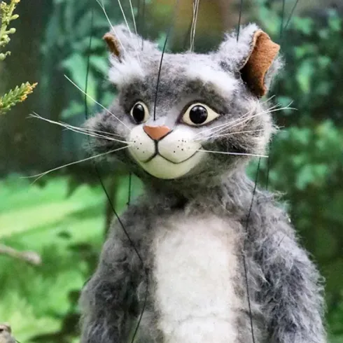 Eine graue Puppenkatze mit großen Augen und einem freundlichen Gesicht steht in einem grünen Wald. Die Katze hat eine weiche Fellstruktur und wirkt lebhaft.
