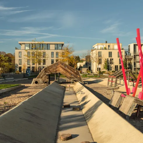 Ein moderner Spielplatz mit bunten Spielgeräten und schattenspendenden Bäumen. Im Hintergrund sind stilvolle Wohngebäude zu sehen.