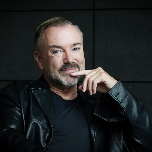 Ein Mann mit einem Bart und einem nachdenklichen Blick sitzt vor einer dunklen Wand. Er trägt eine schwarze Lederjacke und hat seine Hand an sein Kinn gelegt.