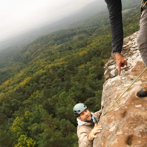 Zwei Kletterer am Felsen in einer grünen, bewaldeten Landschaft. Der eine Kletterer erreicht die Spitze, während der andere ihm Anweisungen gibt.