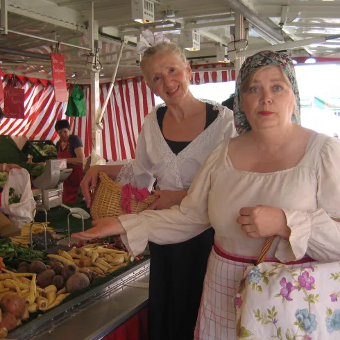 Zwei Frauen auf einem Markt stehen vor einem Tisch mit frischem Gemüse. Im Hintergrund sind weitere Marktstände mit roten und weißen Streifen zu sehen.