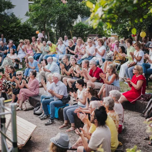 Eine große Menge von Menschen sitzt auf Bänken und applaudiert in einem Garten. Die Atmosphäre ist festlich und fröhlich.