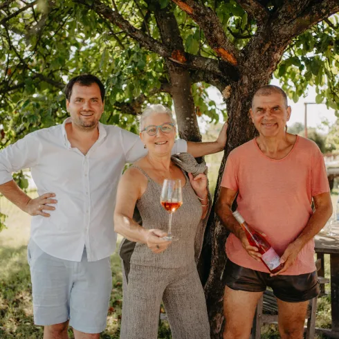 Drei Personen stehen unter einem Baum und lächeln. Sie halten Getränke in der Hand und wirken entspannt.