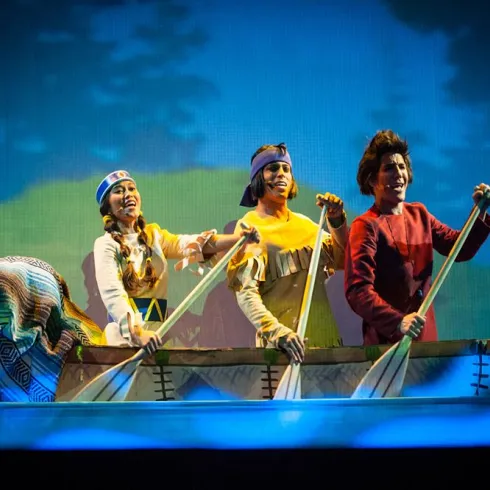 Eine Gruppe von Darstellern in farbenfrohen Kostümen sitzt in einem Boot und rudert. Im Hintergrund ist eine malerische Landschaft mit Bäumen und Blau zu sehen.