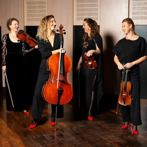 Eine Gruppe von vier Musikerinnen steht in einem modernen Raum. Sie spielen Streichinstrumente und tragen elegante schwarze Kleidung mit roten Schuhen.