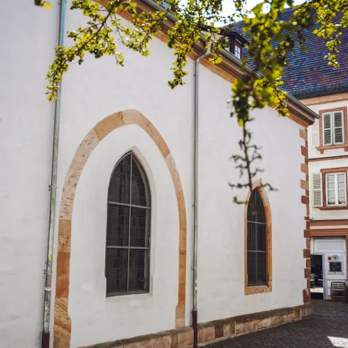 Eine historische Gebäudeansicht mit einem weißen Wand, einem großen, gotischen Fenster und einem Nachbargebäude aus Stein. Die Szene ist von sanften Baumzweigen umrahmt.