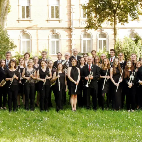 Eine große Gruppe von Musikern steht im Freien auf einer grünen Wiese. Sie halten verschiedene Musikinstrumente und lächeln für das Foto.