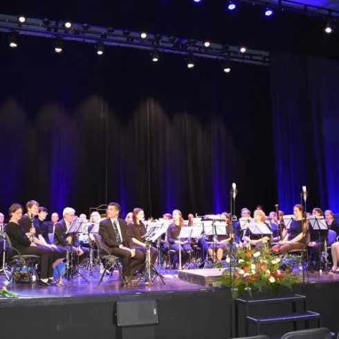Ein Orchester sitzt auf der Bühne mit verschiedenen Instrumenten. Die Beleuchtung ist dunkelblau und es gibt Blumenarrangements im Vordergrund.