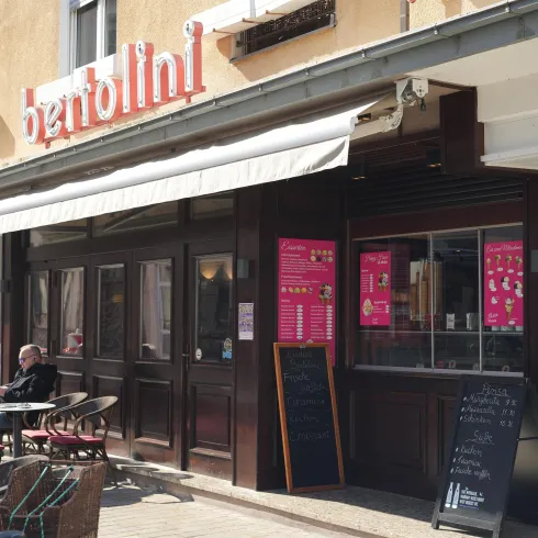 Ein gemütliches Restaurant mit einer einladenden Terrassenfläche. Die Fassade ist in dunklem Holz gehalten und es gibt ein großes Schaufenster mit einer Speisekarte.