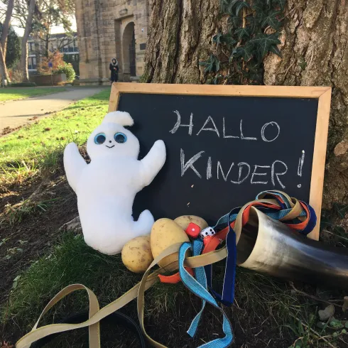 Ein lustiger Geist und eine Tafel mit der Aufschrift „HALLO KINDER!“ stehen neben einigen bunten Gegenständen. Die Szene ist freundlich und einladend, perfekt für Kinder.