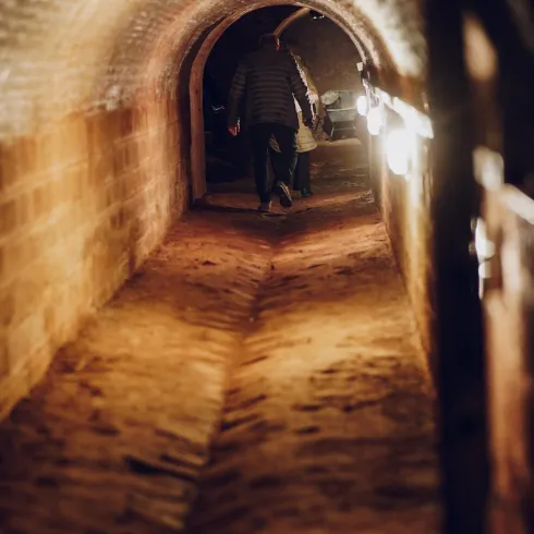 Ein schmaler Tunnel mit beleuchteten Wänden und einem sandigen Boden. Eine Person wandert durch den Tunnel.