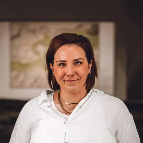 Eine Person mit kurzen, braunen Haaren lächelt in die Kamera. Im Hintergrund ist ein unscharfes Bild zu sehen.