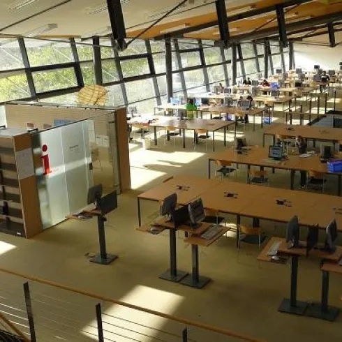 Ein heller, moderner Arbeitsraum mit großen Fenstern und vielen Tischen. In der Mitte stehen Computer und Regale mit Unterlagen.