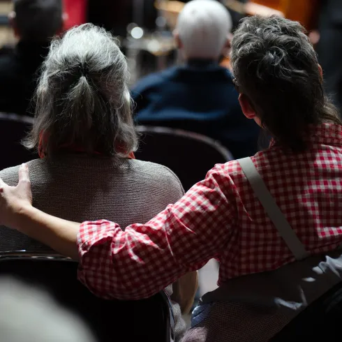 Zwei Menschen sitzen in einem Raum und hören einer Veranstaltung zu. Eine Person umarmt die andere sanft von hinten.
