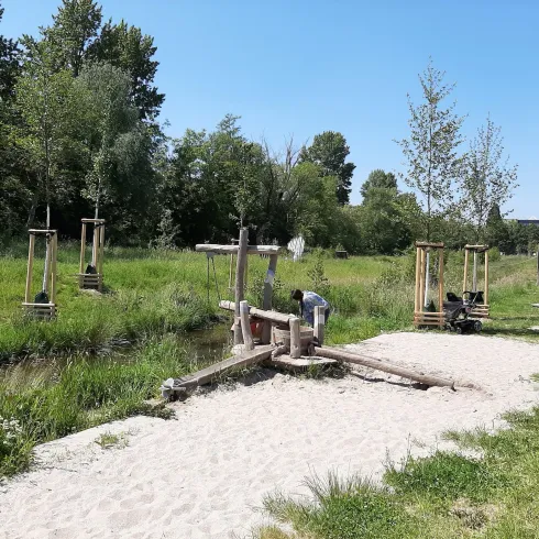 Ein Spielplatz am Wasser mit Holzgeräten und einer sandigen Uferzone. Im Hintergrund sind Bäume und Wiese zu sehen.