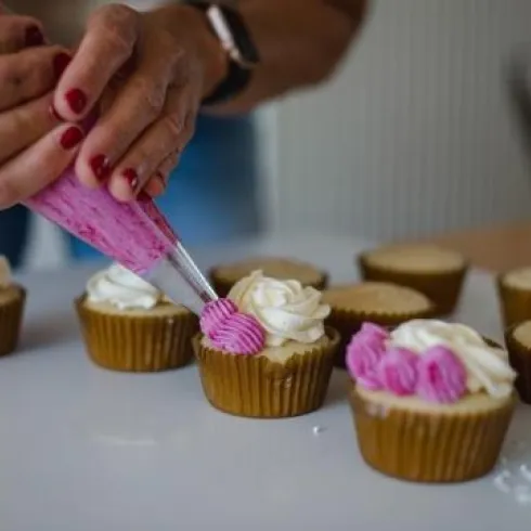 Zwei Hände spritzen rosa und weiße Buttercreme auf frisch gebackene Cupcakes. Im Hintergrund sind weitere Cupcakes zu sehen, die bereits verziert sind.