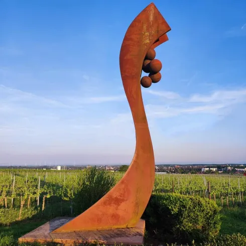 Eine massive, abstrakte Skulptur steht inmitten von Weinreben unter einem klaren blauen Himmel. Die Form ist geschwungen und hat mehrere Kugeln auf der Spitze.