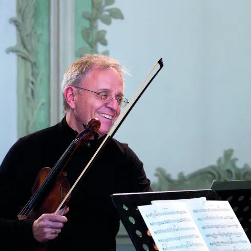 Ein lächelnder Geiger hält seine Violine und steht vor Notenblättern. Der Hintergrund zeigt eine elegante Wandgestaltung.