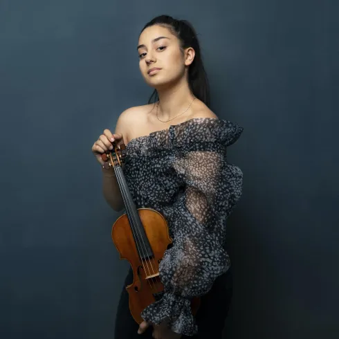 Eine junge Frau steht vor einem blauen Hintergrund und hält eine Violine in der Hand. Sie trägt ein auffälliges, gemustertes Oberteil und hat ihre Haare zurückgebunden.