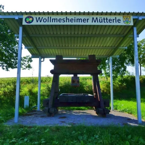 Eine alte Holzpresse steht unter einem überdachten Schutzdach. Im Hintergrund sind grüne Wiesen und Bäume zu sehen.