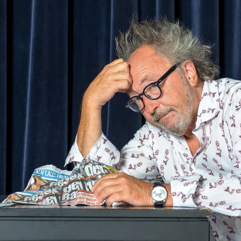 Ein nachdenklicher Mann mit zerzaustem Haar sitzt an einem Tisch und hält eine zusammengeknüllte Zeitung in der Hand. Er sieht besorgt oder frustriert aus.