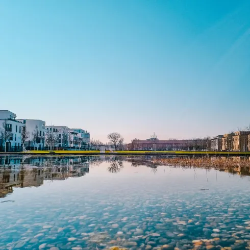 Eine ruhige Szenerie mit einem ruhigen Gewässer, das die umliegenden Gebäude widerspiegelt. Der Himmel ist klar und blau, was eine friedliche Atmosphäre schafft.