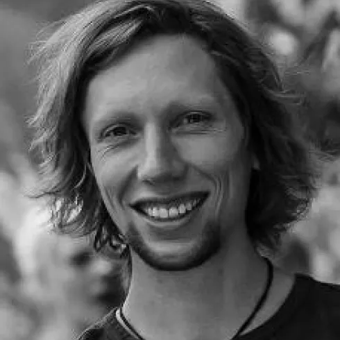 Ein junger Mann mit lockigem Haar und lächelndem Gesicht. Der Hintergrund ist unscharf und zeigt eine natürliche Umgebung.