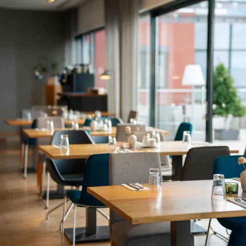 Ein modernes Restaurant mit Holztischen und farbigen Stühlen. Große Fenster sorgen für viel Tageslicht und Blick auf die Terrasse.