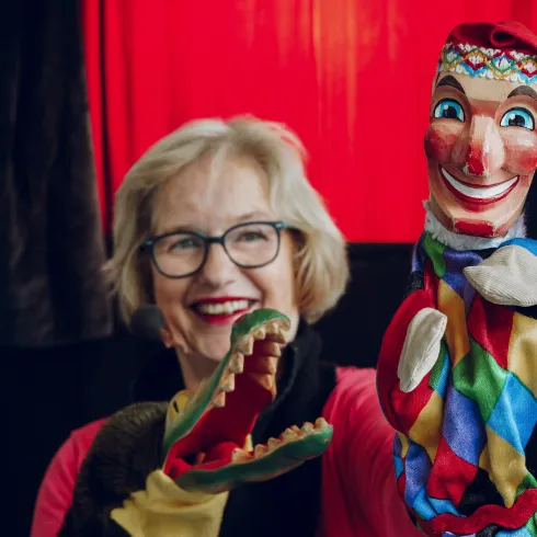 Eine Frau lächelt glücklich und hält zwei Handpuppen in den Händen. Im Hintergrund sind rote Vorhänge zu sehen.