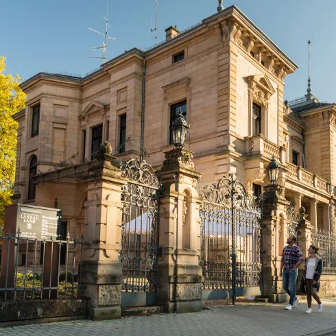 Ein beeindruckendes historisches Gebäude mit kunstvollem Gitterzaun. Im Vordergrund gehen zwei Personen spazieren.