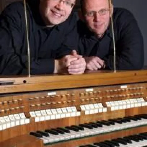 Zwei Männer stehen lächelnd hinter einer Orgel. Die Orgel hat viele Tasten und ist aus Holz gefertigt.